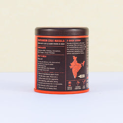 Side view of an orange and black tin of Amala Chai Cardamom Chai Masala showing ingredients, brewing instructions, and information about its origin in Kerala, India.