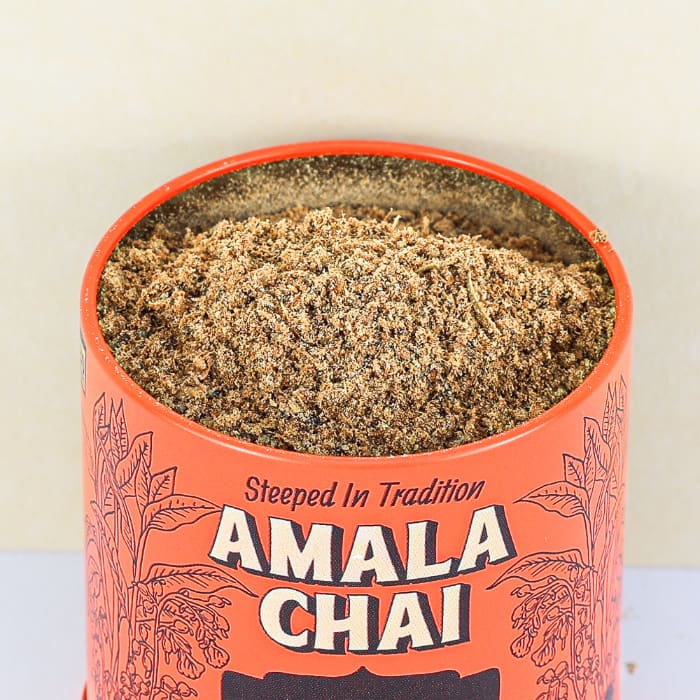 Close-up, top-down view of an open orange tin of Amala Chai Cardamom Chai Masala, revealing the loose-leaf tea and spice blend inside.