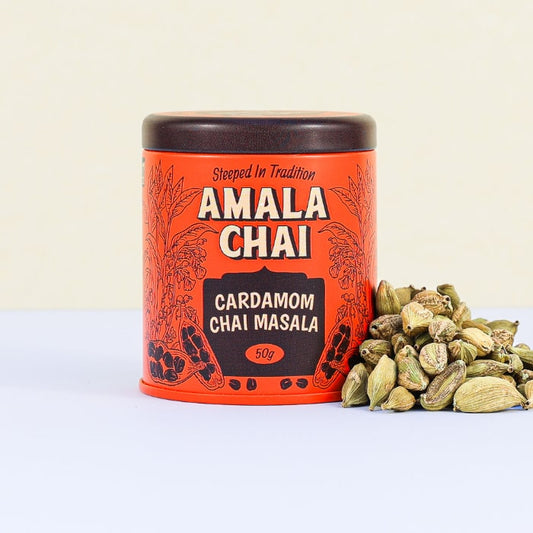 Orange tin of Amala Chai Cardamom Chai Masala with black lettering and illustrations, next to a pile of green cardamom pods.