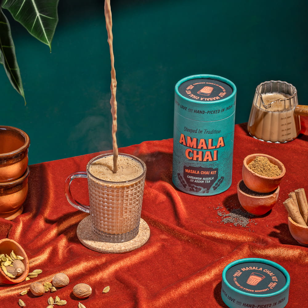 A teal cylindrical tin of Amala Chai Masala Chai Kit on a red surface, with a stream of chai being poured into a glass mug, surrounded by spices like cardamom, nutmeg, and cinnamon sticks, along with small bowls of tea powder.