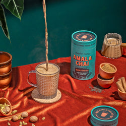 A teal cylindrical tin of Amala Chai Masala Chai Kit on a red surface, with a stream of chai being poured into a glass mug, surrounded by spices like cardamom, nutmeg, and cinnamon sticks, along with small bowls of tea powder.