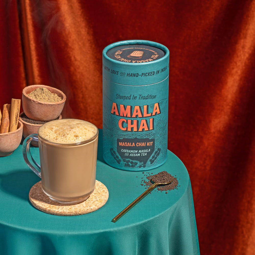 Teal cylindrical tin of Amala Chai Masala Chai Kit on a teal surface next to a glass mug of frothy chai on a cork coaster, with a spoon and loose tea beside it, and small bowls of spices in the background.
