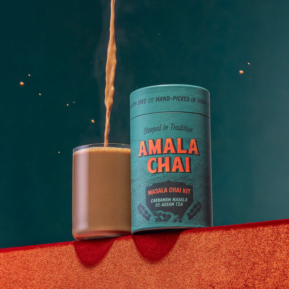 A teal cylindrical tin of Amala Chai Masala Chai Kit on a textured orange surface, with a stream of chai being poured into a clear glass mug against a dark teal background.