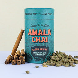 Teal cylindrical tin of Amala Chai Masala Chai Kit with orange lettering, featuring cardamom masala and Assam tea, surrounded by cinnamon sticks and green cardamom pods.