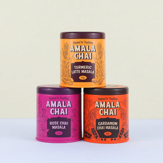 A stack of three Amala Chai tins: a golden orange tin of Turmeric Latte Masala on top, a magenta tin of Rose Chai Masala in the middle, and an orange tin of Cardamom Chai Masala at the bottom.