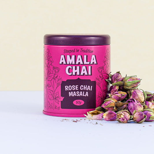 Magenta tin of Amala Chai Rose Chai Masala with dark purple lettering and rose illustrations, next to a pile of dried rosebuds.