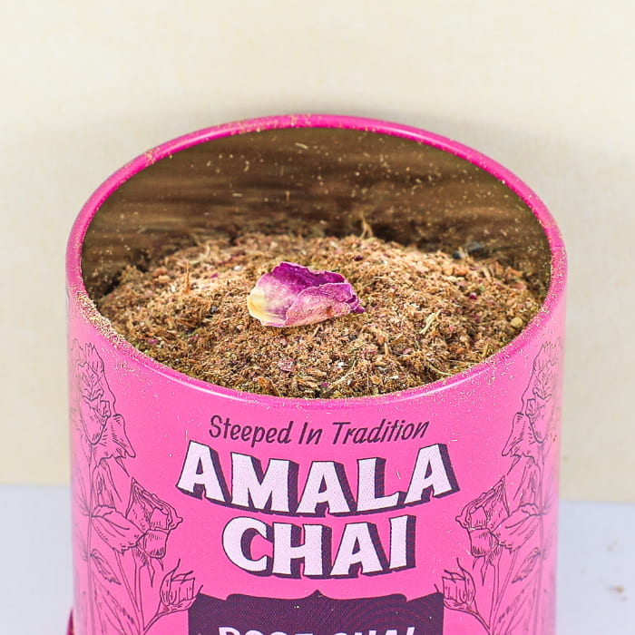 Close-up, top-down view of an open magenta tin of Amala Chai Rose Chai Masala, revealing the loose-leaf tea and spice blend with a single rose petal on top.