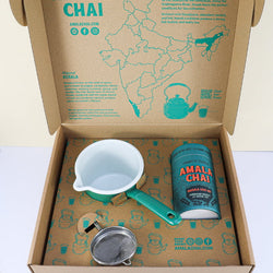 An open cardboard gift box of Amala Chai Masala Chai Kit, revealing a teal cylindrical tin, a small green and white enamel saucepan, and a stainless steel tea strainer, all nestled on patterned card featuring teapots and a map of India.