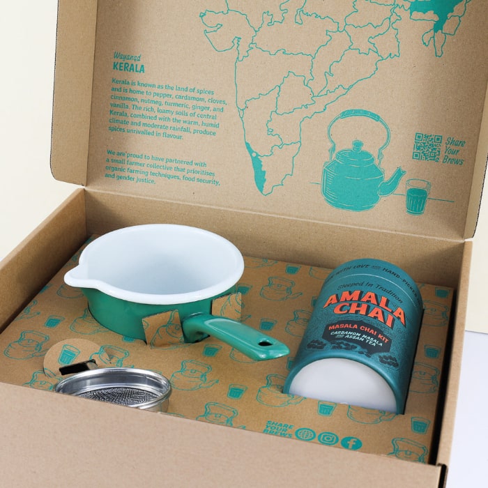 An open cardboard gift box of Amala Chai Masala Chai Kit photographed at an angle, showing a teal cylindrical tin, a small green and white enamel saucepan secured with cardboard, and a stainless steel tea strainer, arranged on patterned paper with teapots and a map of India highlighting Kerala.