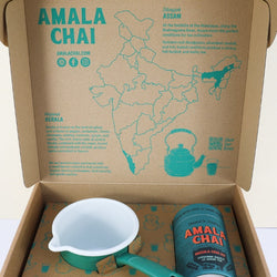 A more close up image of an open cardboard gift box of Amala Chai Masala Chai Kit, featuring a teal cylindrical tin, a small green and white enamel saucepan held in place by cardboard, and the inside lid printed with information about Assam and Kerala, alongside a map of India.