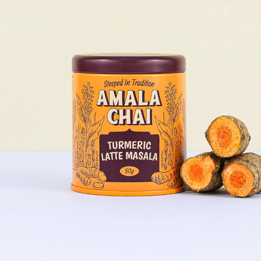 Golden orange tin of Amala Chai Turmeric Latte Masala with dark brown lettering and turmeric root illustrations, next to three cut turmeric roots.