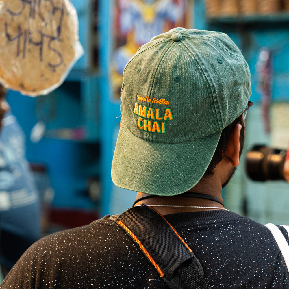 the back of a mans head who's wearing an Amala Chai branded cap. The cap is dark green dyed cotton with a washed out effect.