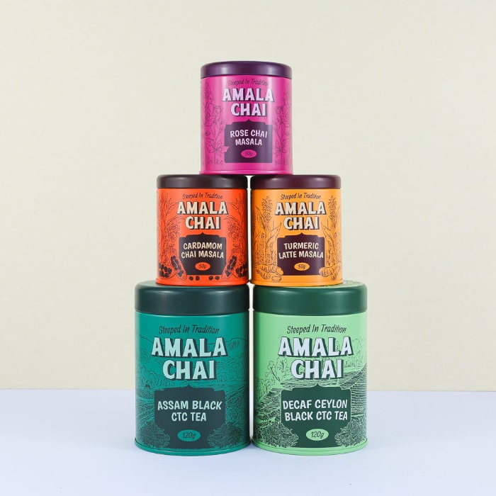 A stack of five Amala Chai tins: a magenta tin of Rose Chai Masala on top, followed by an orange tin of Cardamom Chai Masala and a golden orange tin of Turmeric Latte Masala in the middle row, and a teal tin of Assam Black CTC Tea and a light green tin of Decaf Ceylon Black CTC Tea on the bottom.
